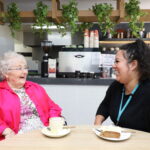 Female resident having a coffee, chatting with one of the carers, in the cafe at Mercy Place Parkville.