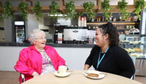 Female resident having a coffee, chatting with one of the carers, in the cafe at Mercy Place Parkville.